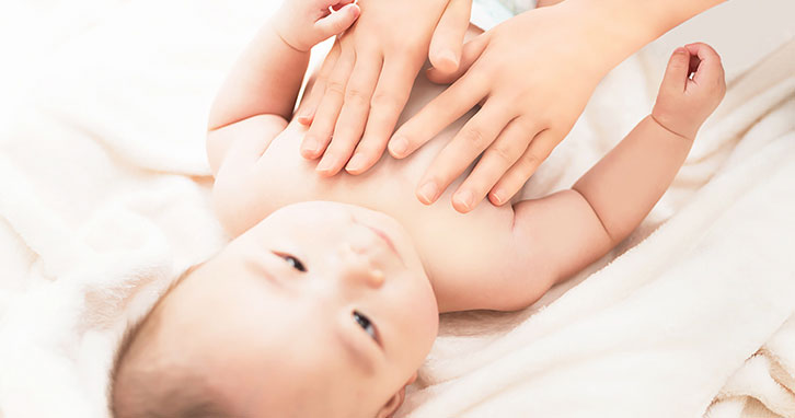 白い布の上に寝ている赤ちゃんと、赤ちゃんのお肌に手のひらをあてて赤ちゃんスキンケアをしているお母さん。穏やかなスキンケア タイムを過ごす親子の画像。